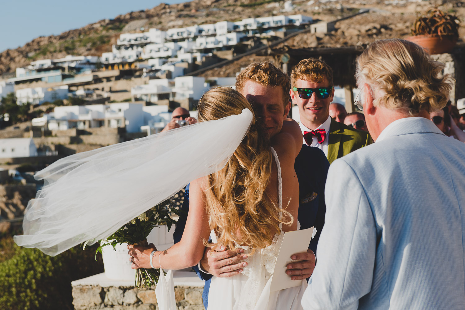 Wedding photographer Mykonos: bride and groom hug with her veil in the wind during their Mykonos wedding.