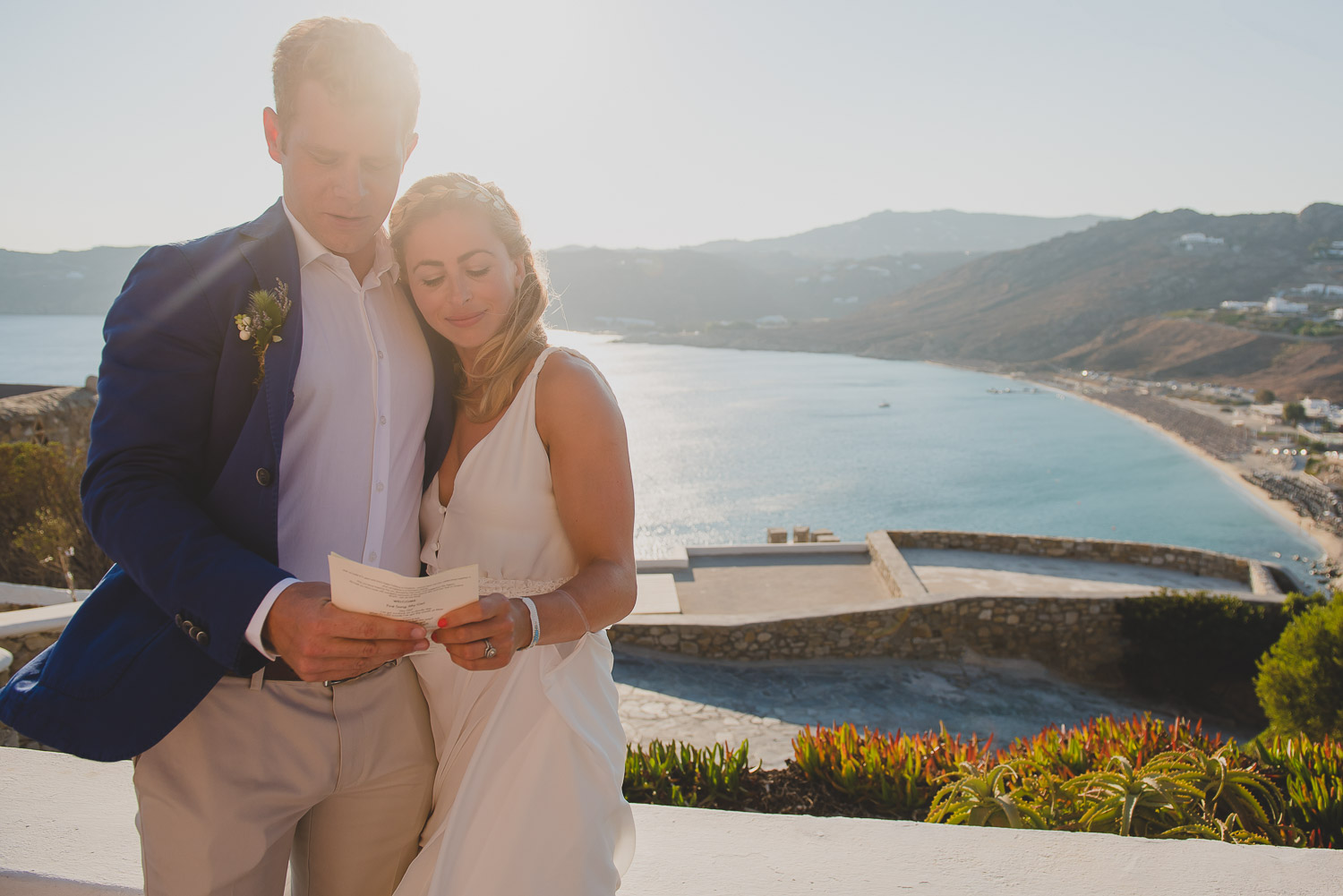 Wedding photographer Mykonos: bride and groom backlit and embraced during Mykonos wedding ceremony with beach views in the background.