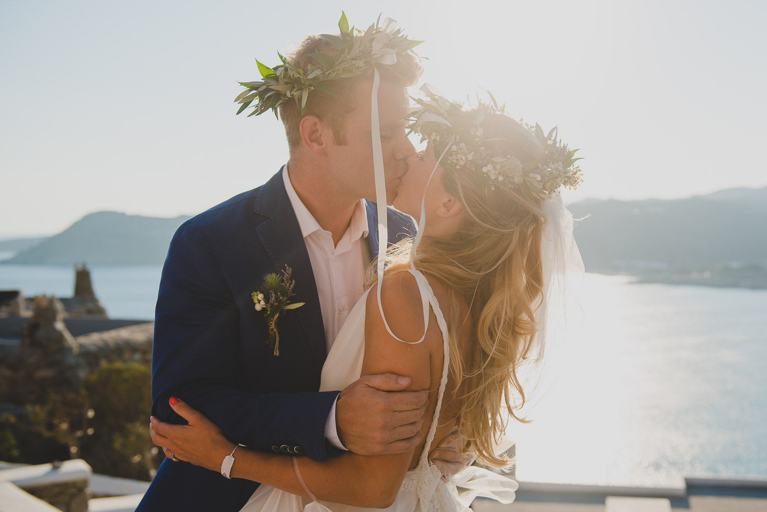 Wedding photographer Mykonos:  newlyweds kiss with olive branch wedding crowns on their heads backlit by the sun after Mykonos wedding ceremony.