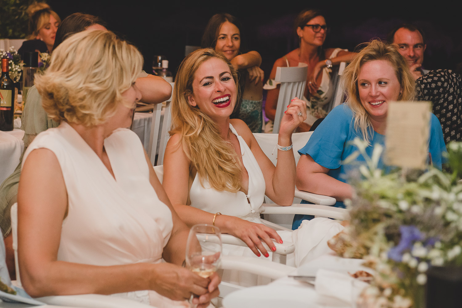Wedding photographer Mykonos: bride and friends laughing during speeches at Elia for Mykonos wedding reception.