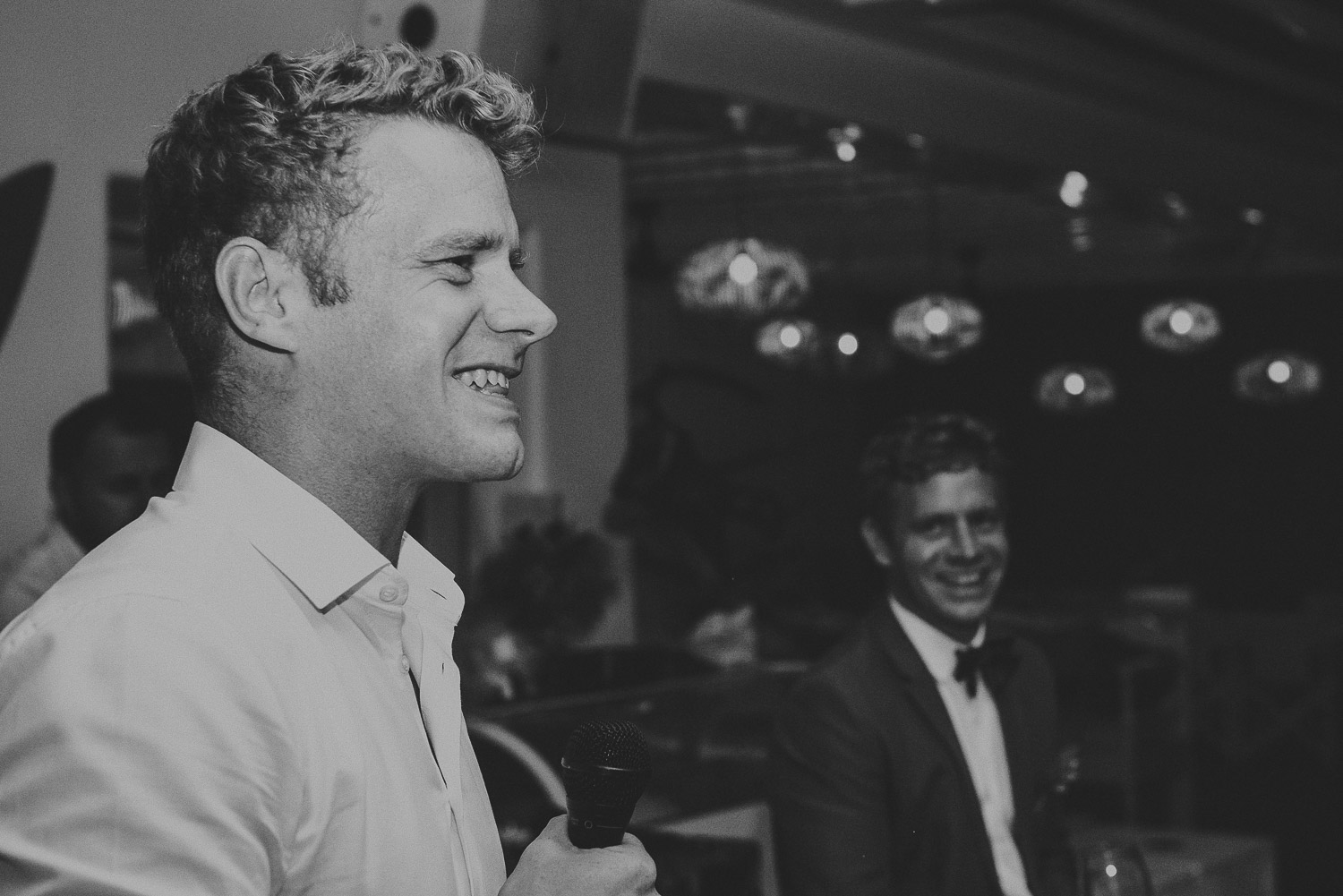 Wedding photographer Mykonos: black and white photo of the groom laughing at Elia during Mykonos wedding speeches.