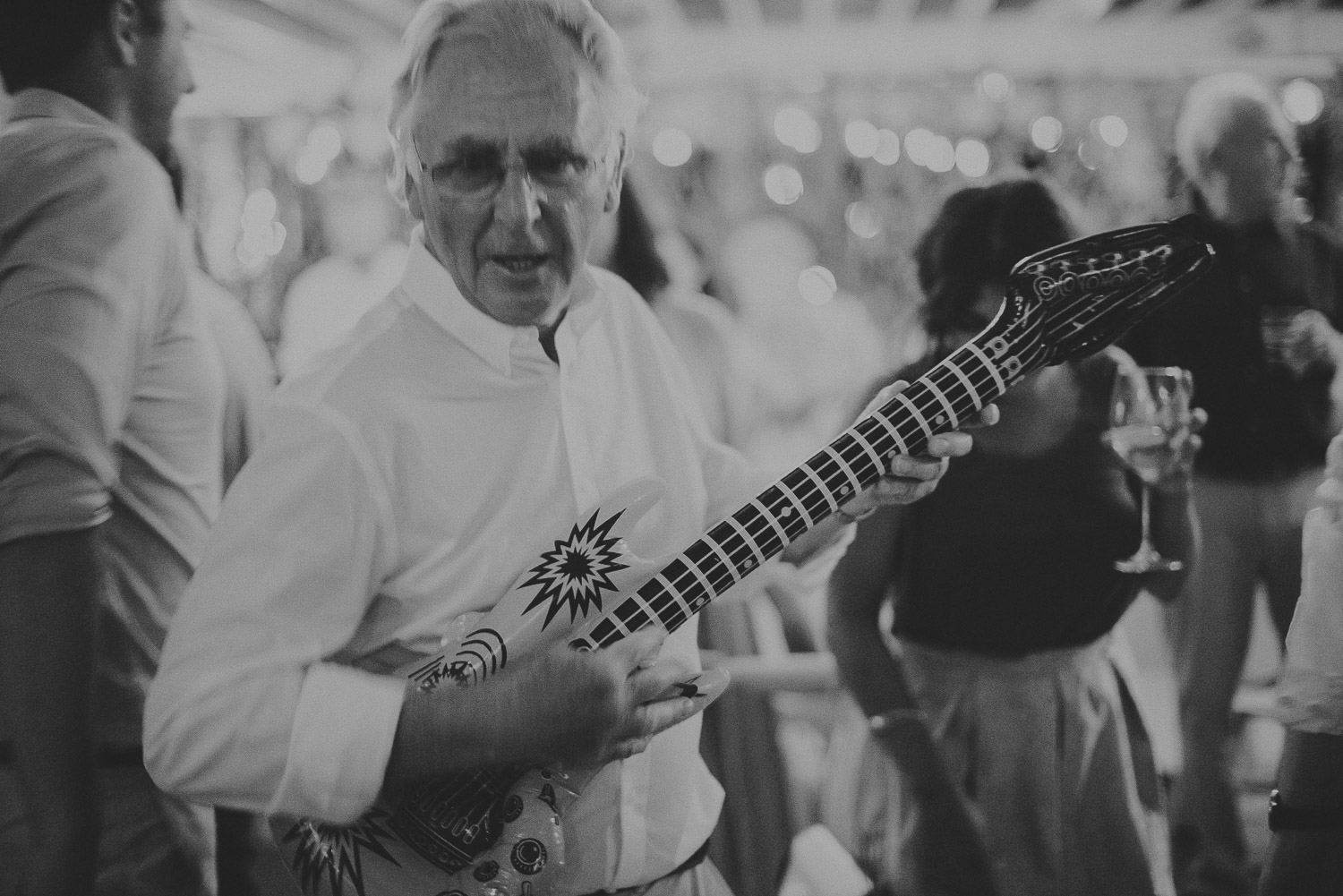 Wedding photographer Mykonos: black and white photo of father of the bride with inflatable guitar at Elia during Mykonos wedding.