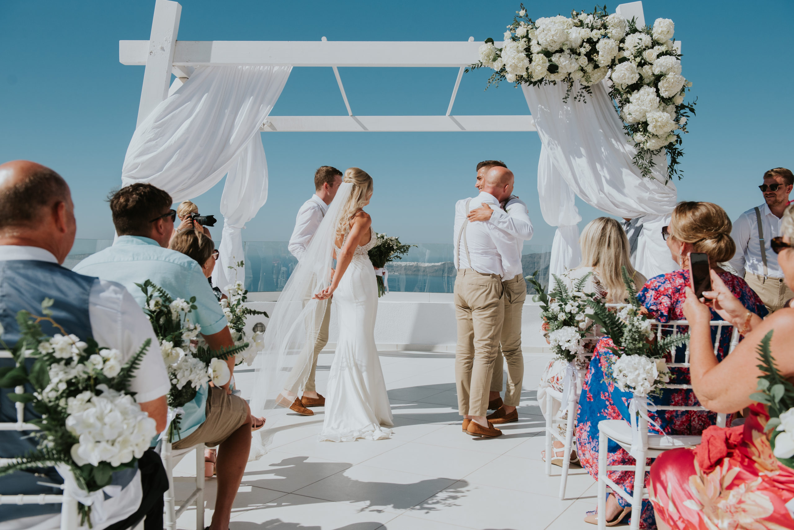 Wedding photographer Santorini: the groom hugs stepfather of the bride in front of gazebo as brides moves her veil by Ben and Vesna.
