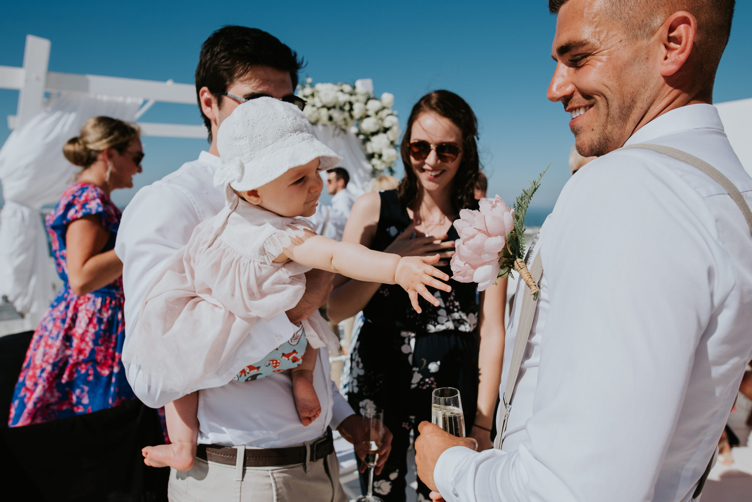 Wedding photographer Santorini:a cute baby reaches for groom's buttoner as he smiles by Ben and Vesna.