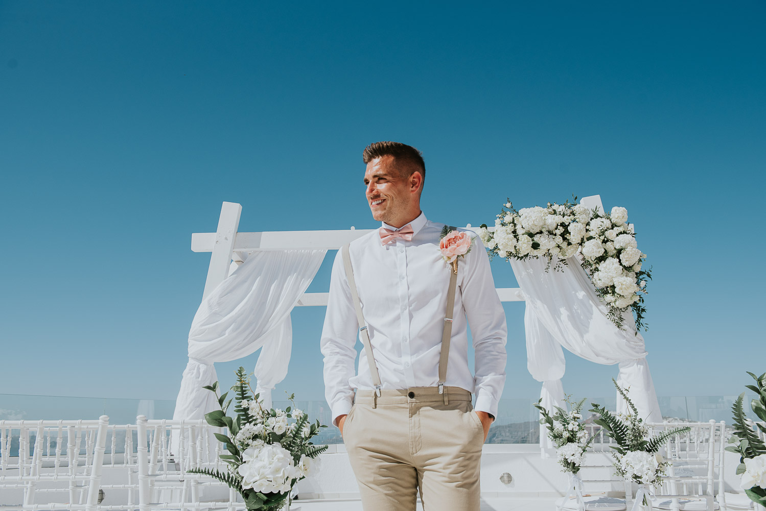 Wedding photographer Santorini: groom walking along the aisle with gazebo covered in flowers in the background by Ben and Vesna.