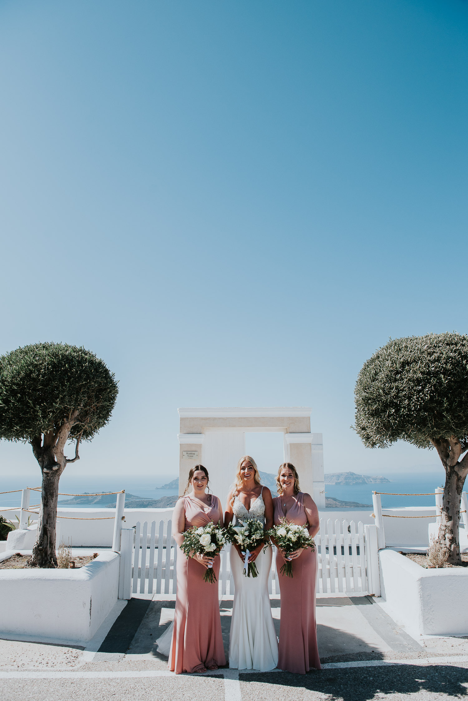Wedding photographer Santorini: bride and her bridesmaids in front of the gate with trees around by Ben and Vesna.