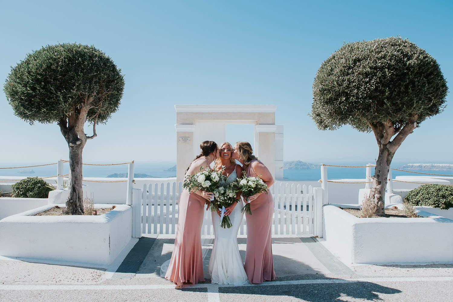 Wedding photographer Santorini: bride gets a kiss on her cheeks by her bridesmaids in front of the gate with trees around by Ben and Vesna.