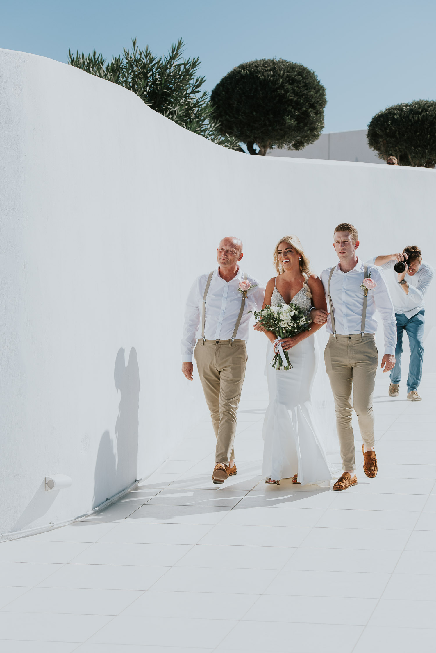 Wedding photographer Santorini: bride smiling walking to the groom with her brother and stepdad by Ben and Vesna.