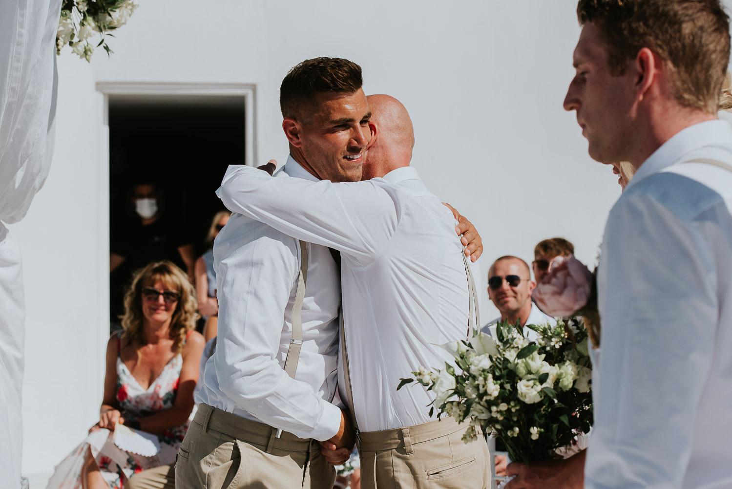 Wedding photographer Santorini: groom and bride's stepdad hug with guests around by Ben and Vesna.