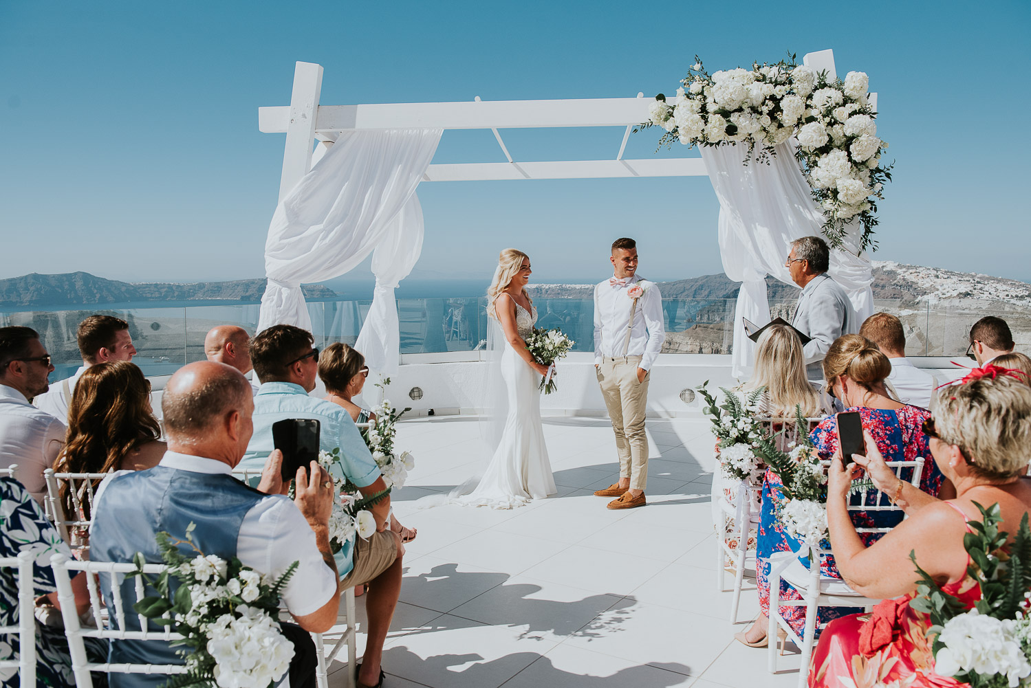 Wedding photographer Santorini: panoramic photo of bride and groom standing in front of gazebo with guests in foreground by Ben and Vesna.
