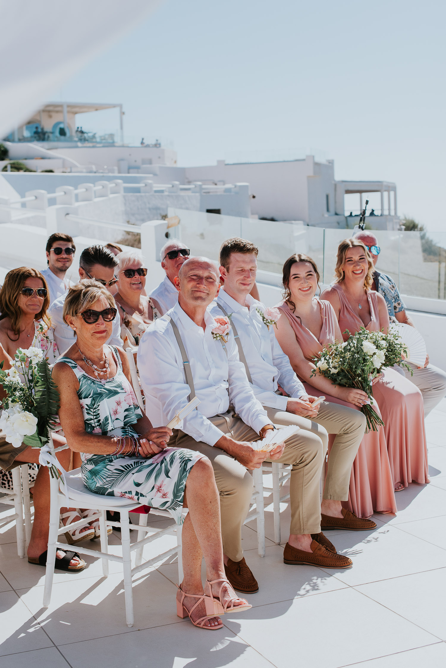 Wedding photographer Santorini: guests sat on the ceremony terrace with bridesmaids smiling in the sunshine by Ben and Vesna.