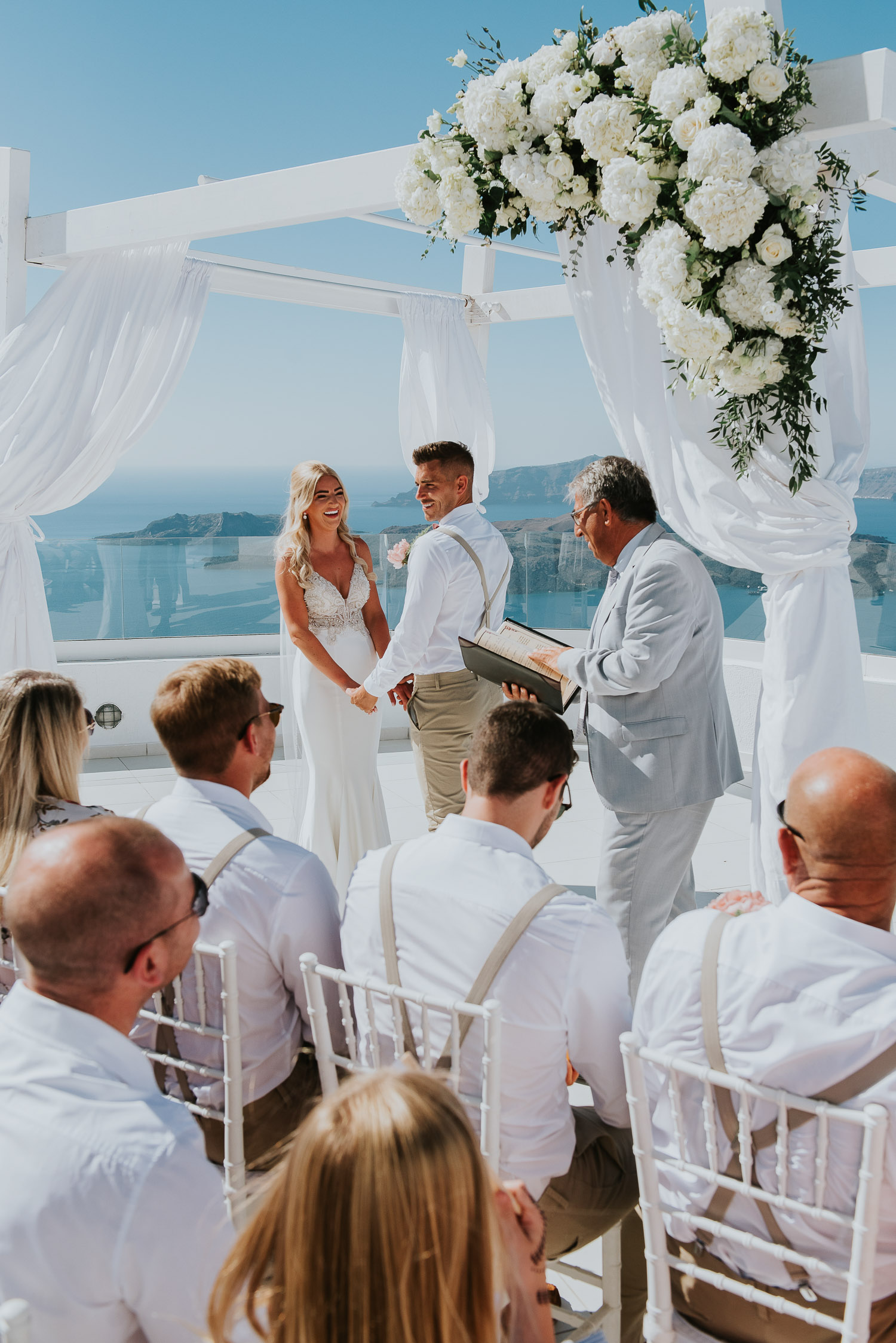 Wedding photographer Santorini: bride and groom standing holding hands in front of gazebo photographed over their guests by Ben and Vesna.
