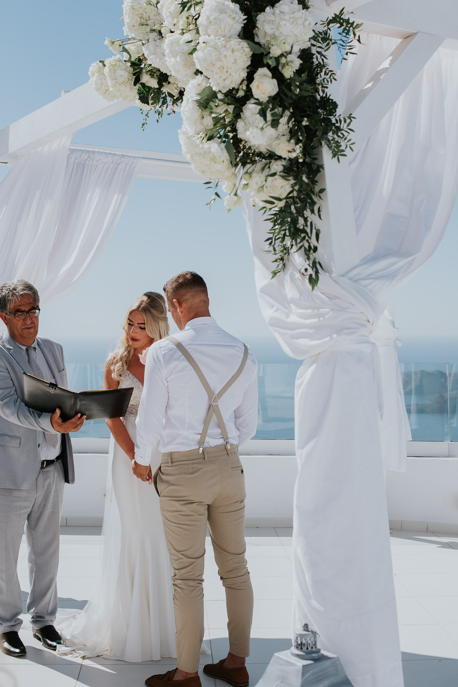 Wedding photographer Santorini: bride and groom holding hands as she reads vows  in front of gazebo by Ben and Vesna.