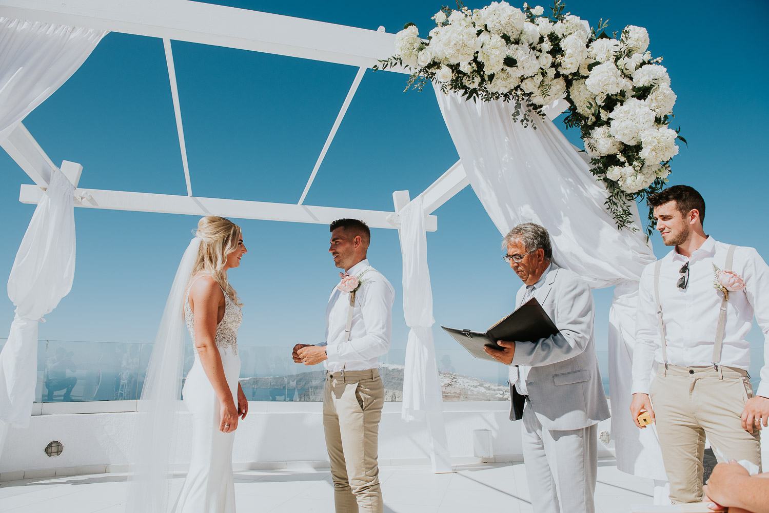 Wedding photographer Santorini: groom holding a ring looking at his bride surrounded by celebrant and the best man by Ben and Vesna.