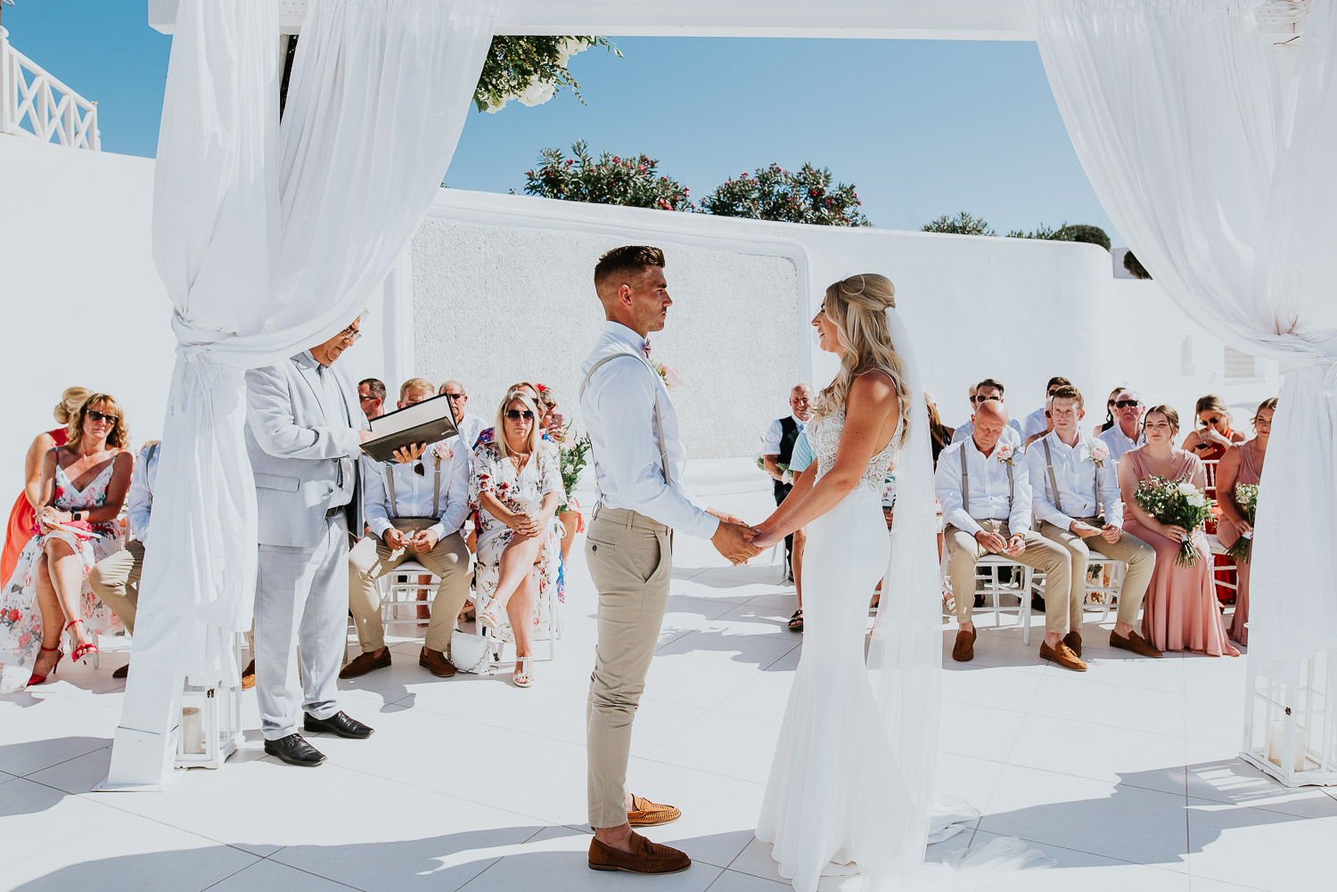 Wedding photographer Santorini: bride and groom holding hands under the gazebo with the guests sat in the background.