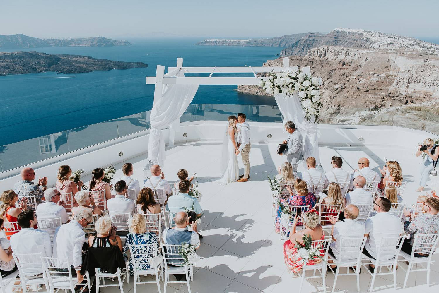 Wedding photographer Santorini: panoramic photo of bride and groom kissing under the gazebo with caldera and the sea in the background by Ben and Vesna.