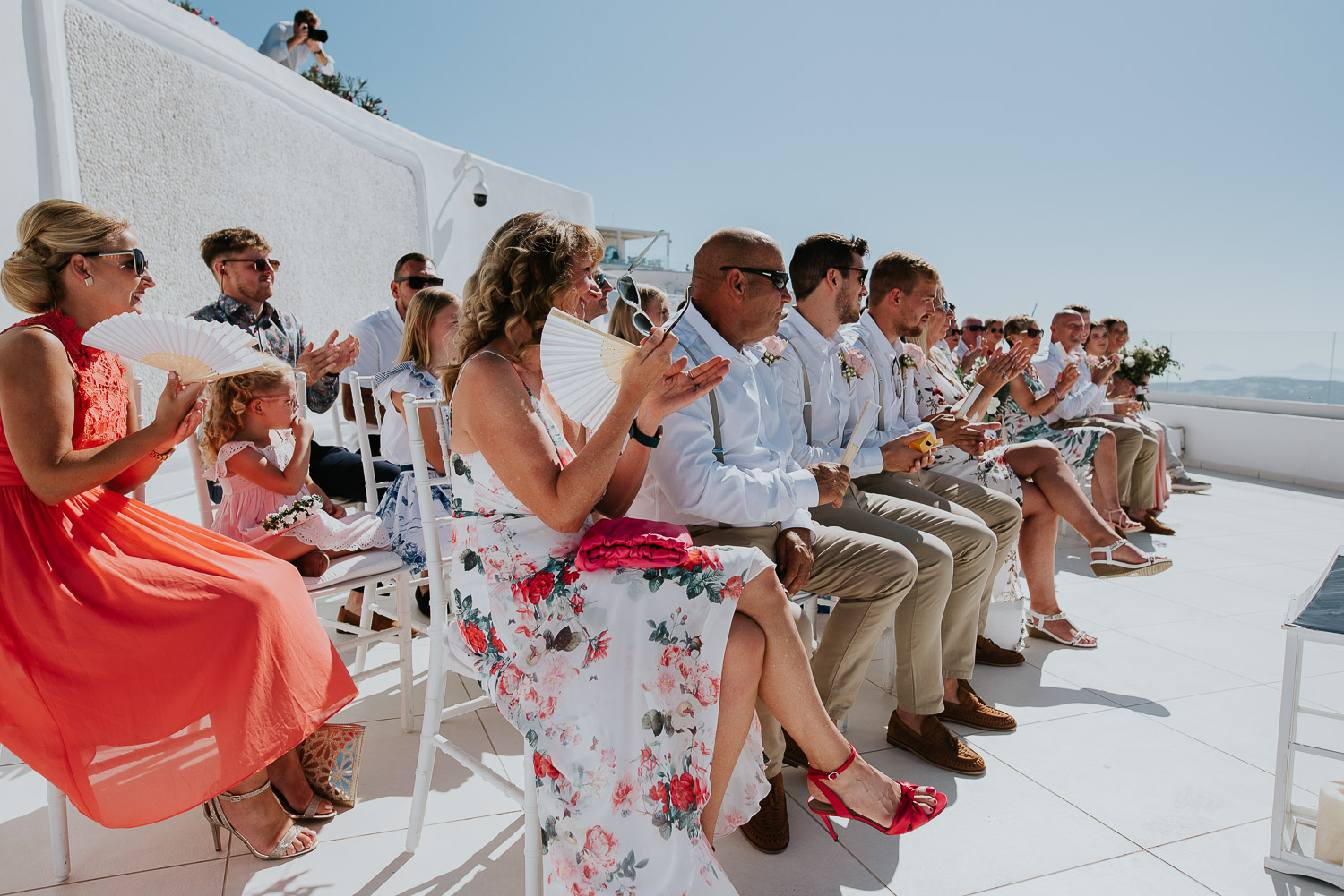 Wedding photographer Santorini: guests clap sat at the ceremony terrace basked in the summer sun by Ben and Vesna.