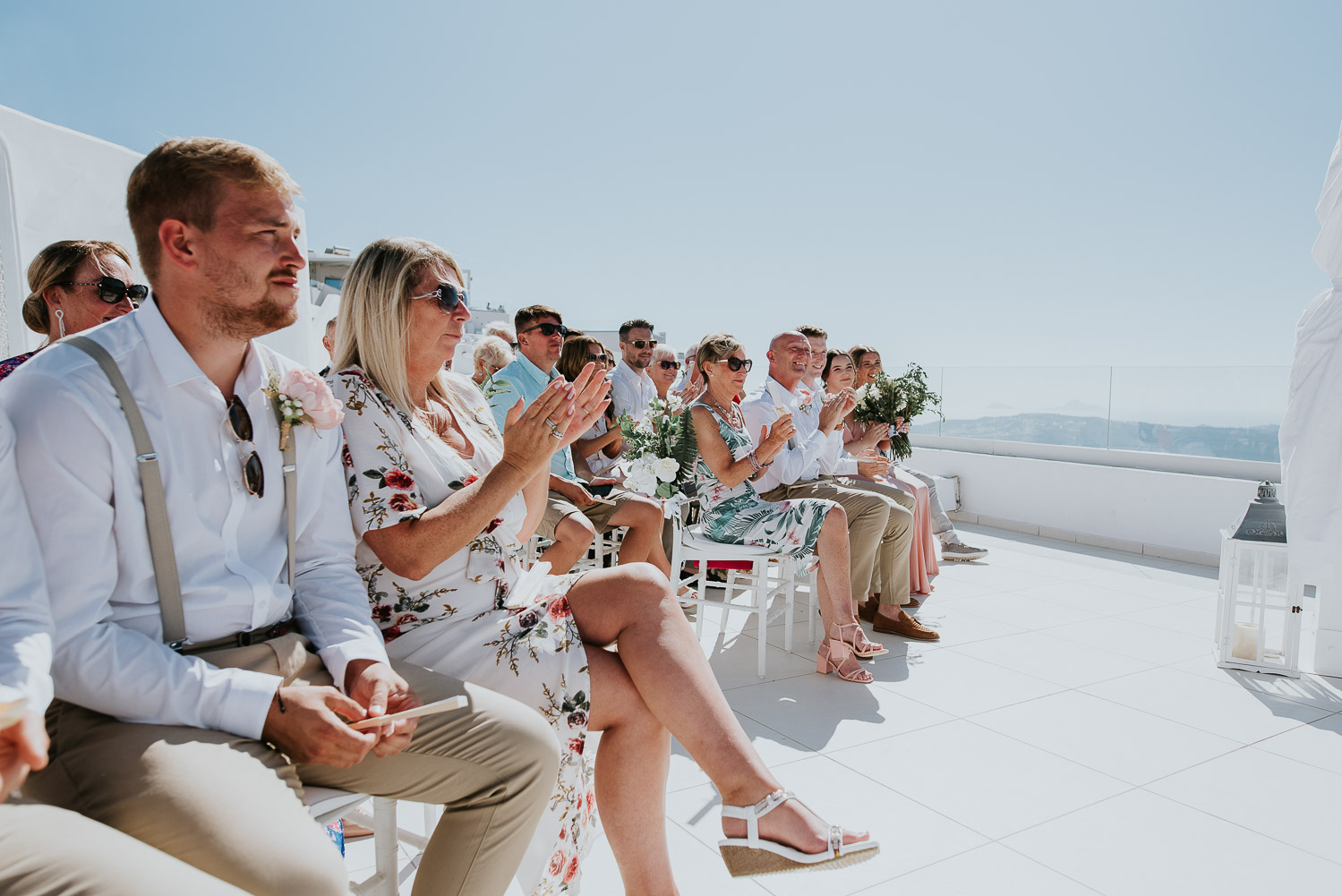 Wedding photographer Santorini: close up pf the guests clapping sat at the ceremony terrace basked in the summer sun by Ben and Vesna.