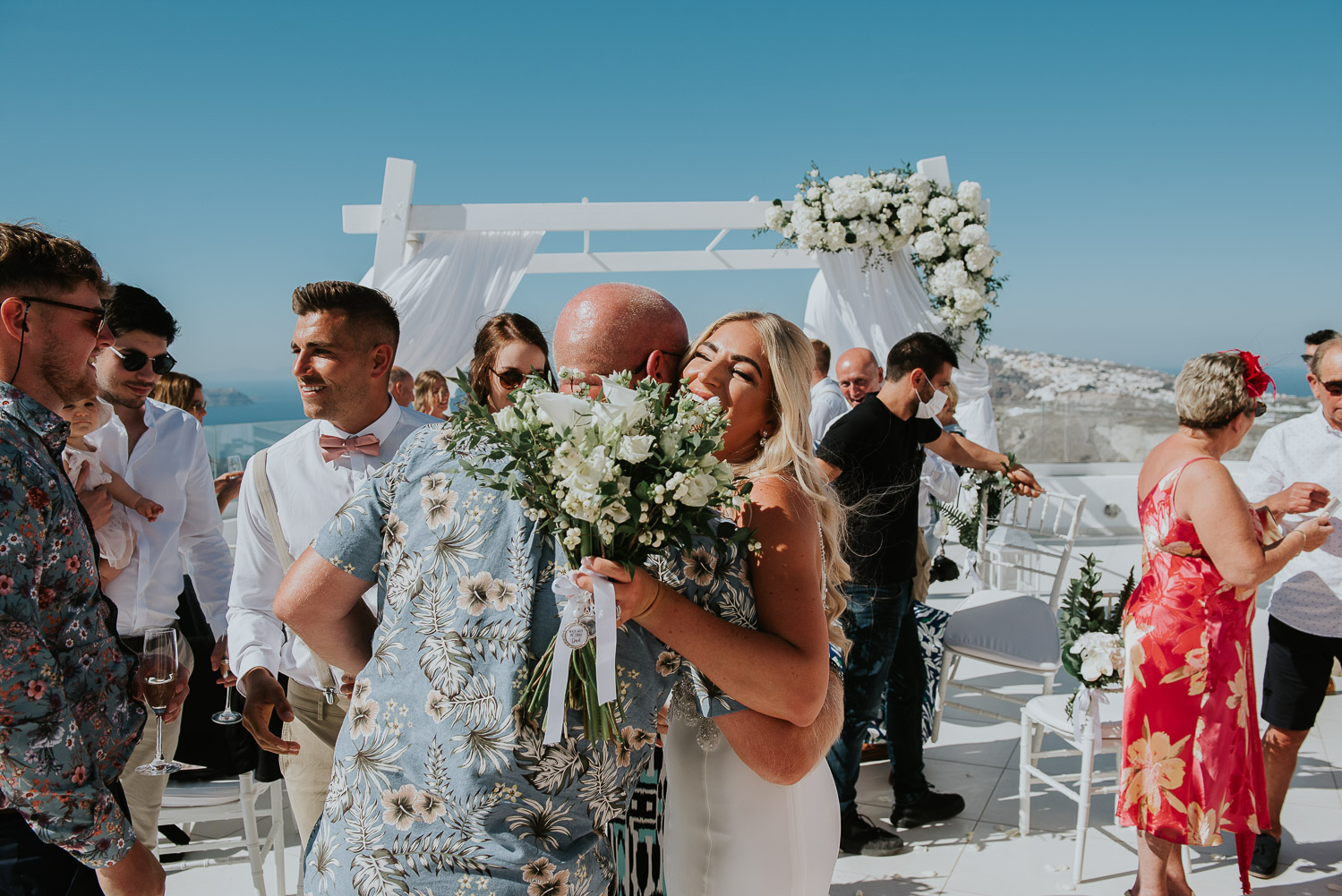 Wedding photographer Santorini: bride smiling hugging one of the guests with groom next to her  by Ben and Vesna.
