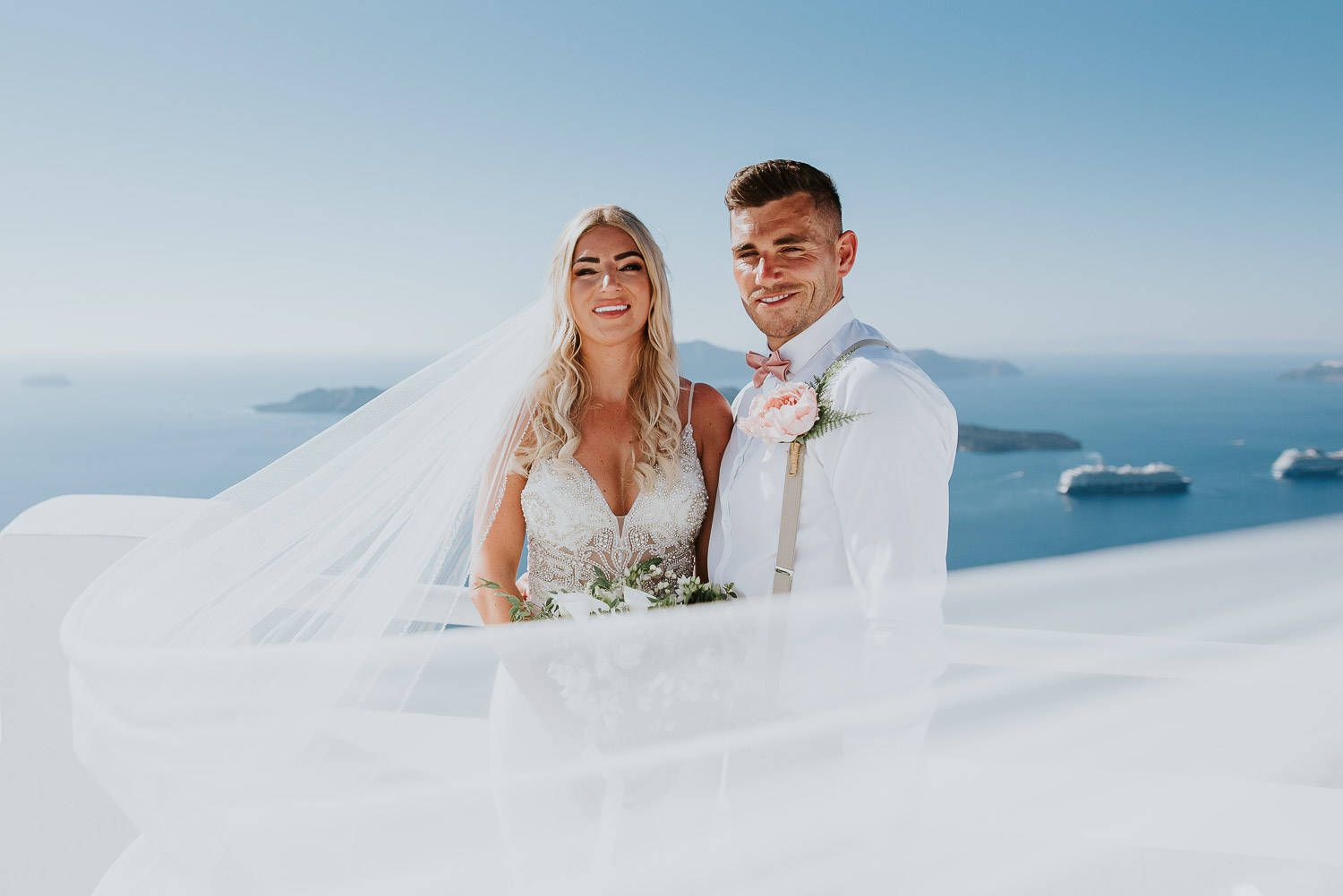 Wedding photographer Santorini: close up of bride and groom engulfed in the veil looking at the camera with the sea in the background by Ben and Vesna.