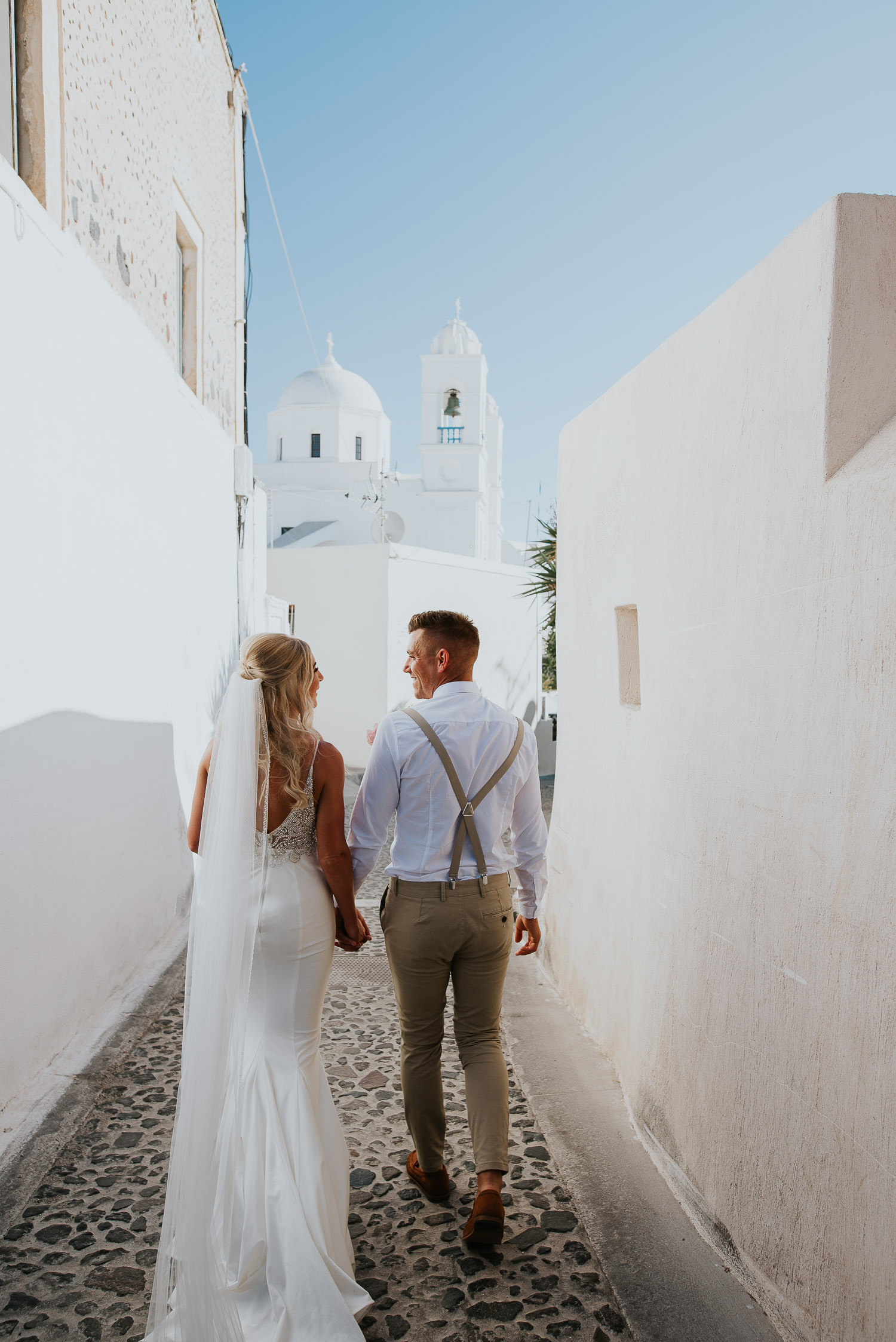 Wedding photographer Santorini: bride and groom walking on a cobbled stone lane with the church in the background by Ben and Vesna.