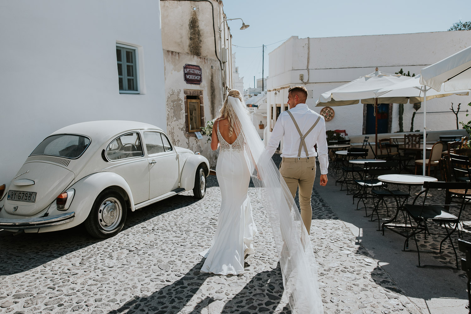 Wedding photographer Santorini: bride and groom crossing a cobbled stone square with old beetle car parked by Ben and Vesna.