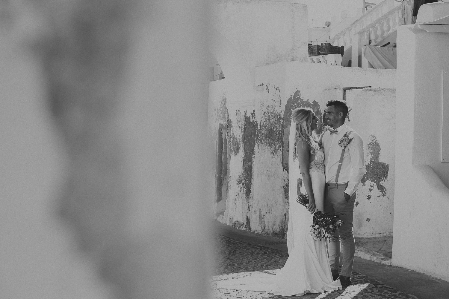 Wedding photographer Santorini: black and white photo of bride and groom against rustic walls standing on cobbled stone street by Ben and Vesna.