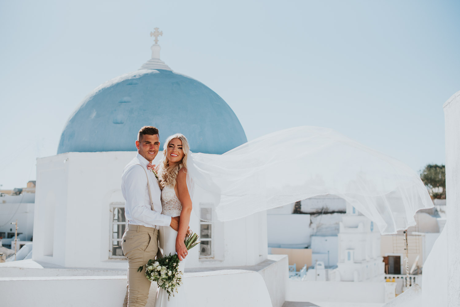 Wedding photographer Santorini: bride and groom looking at the camera with the blue dome behind them and the veil in the wind by Ben and Vesna.