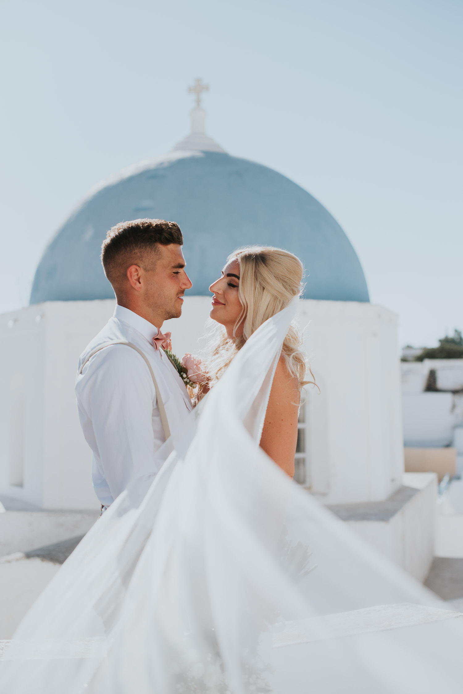Wedding photographer Santorini: close up of bride and groom looking at each other with the blue dome behind and the veil engulfing them by Ben and Vesna.