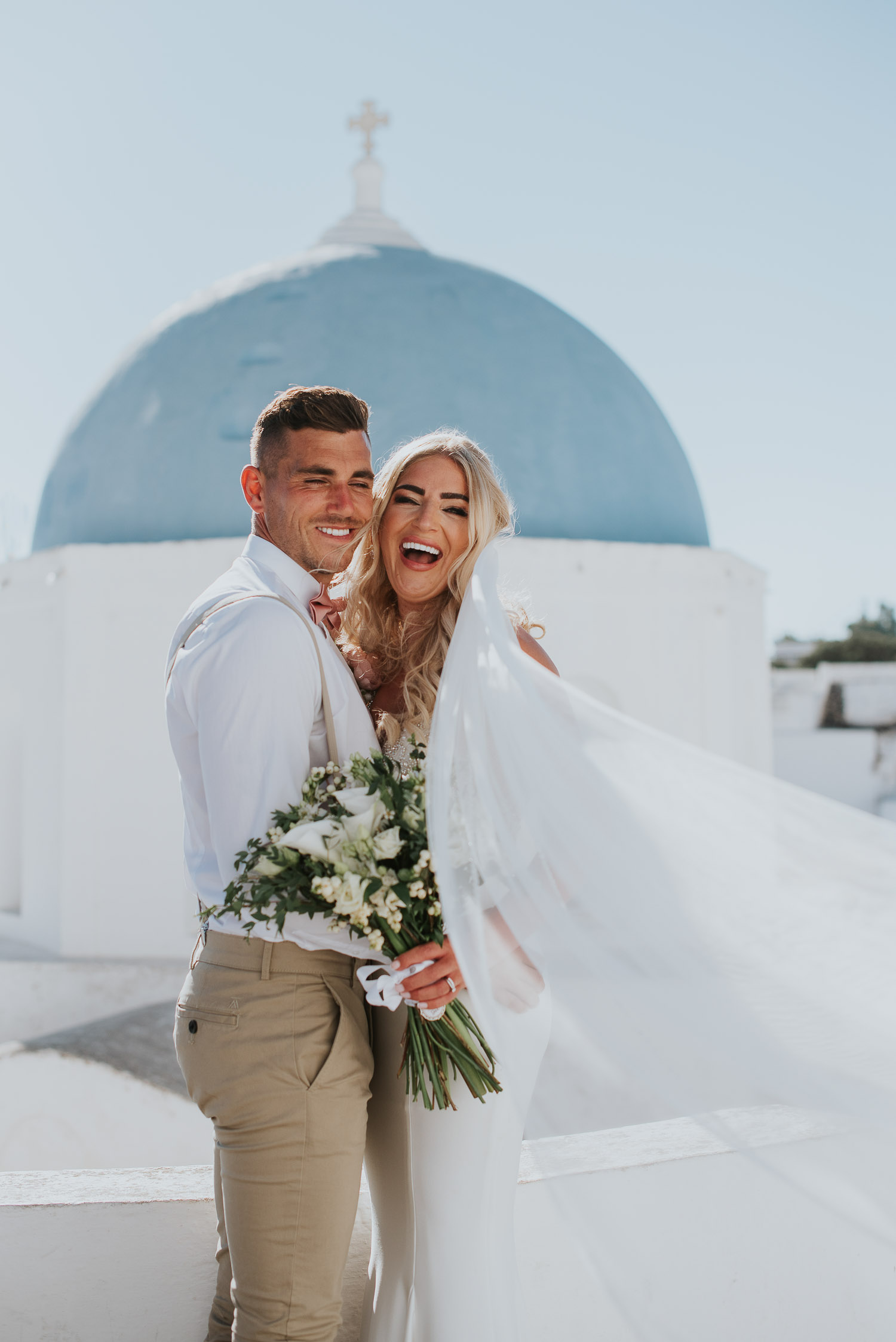 Wedding photographer Santorini captures bride and groom laughing with blue dome behind and the veil engulfing them. Pyrgos restaurant wedding by Ben and Vesna.