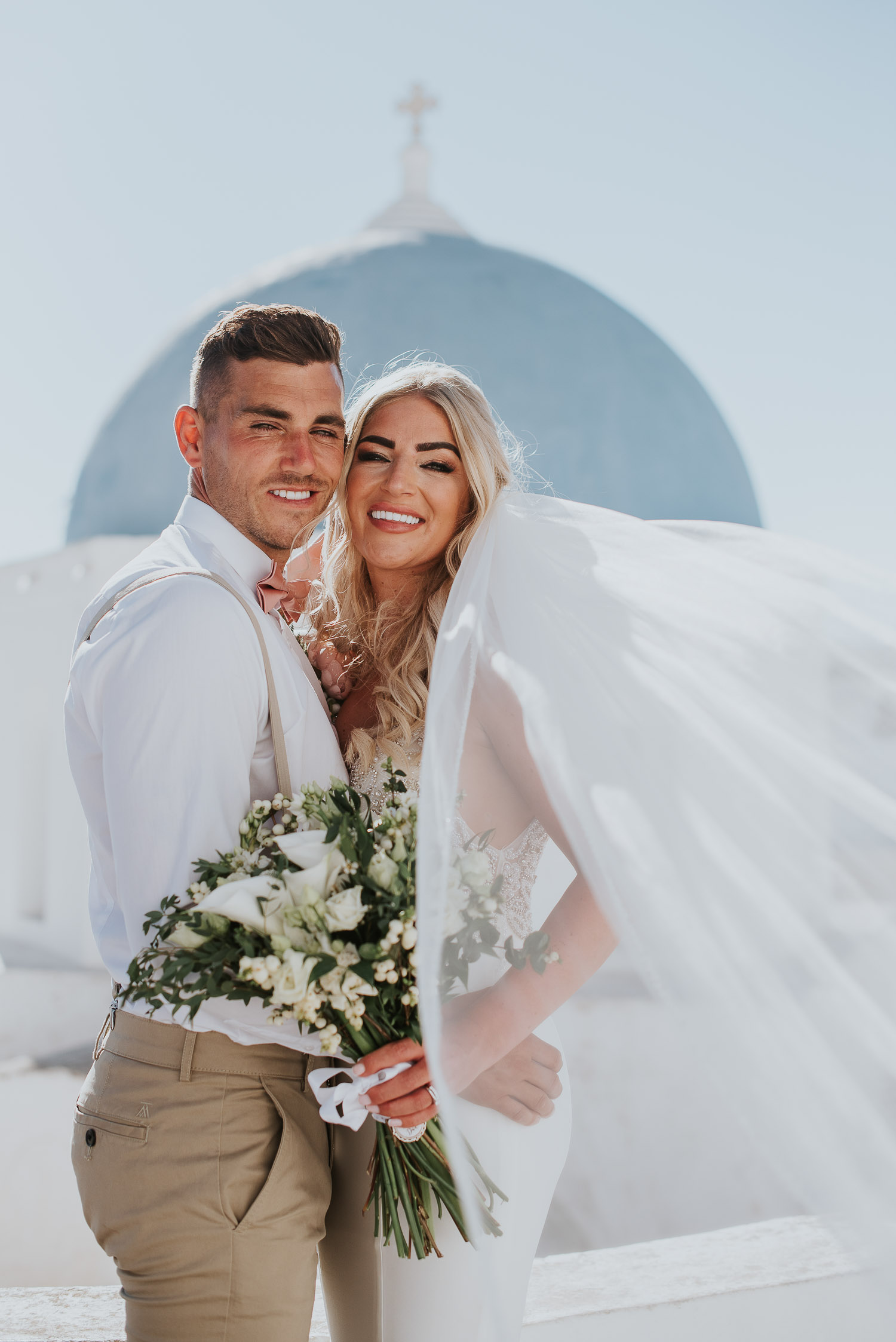 Wedding photographer Santorini: close up of bride and groom with the blue dome behind and her veil them by Ben and Vesna.
