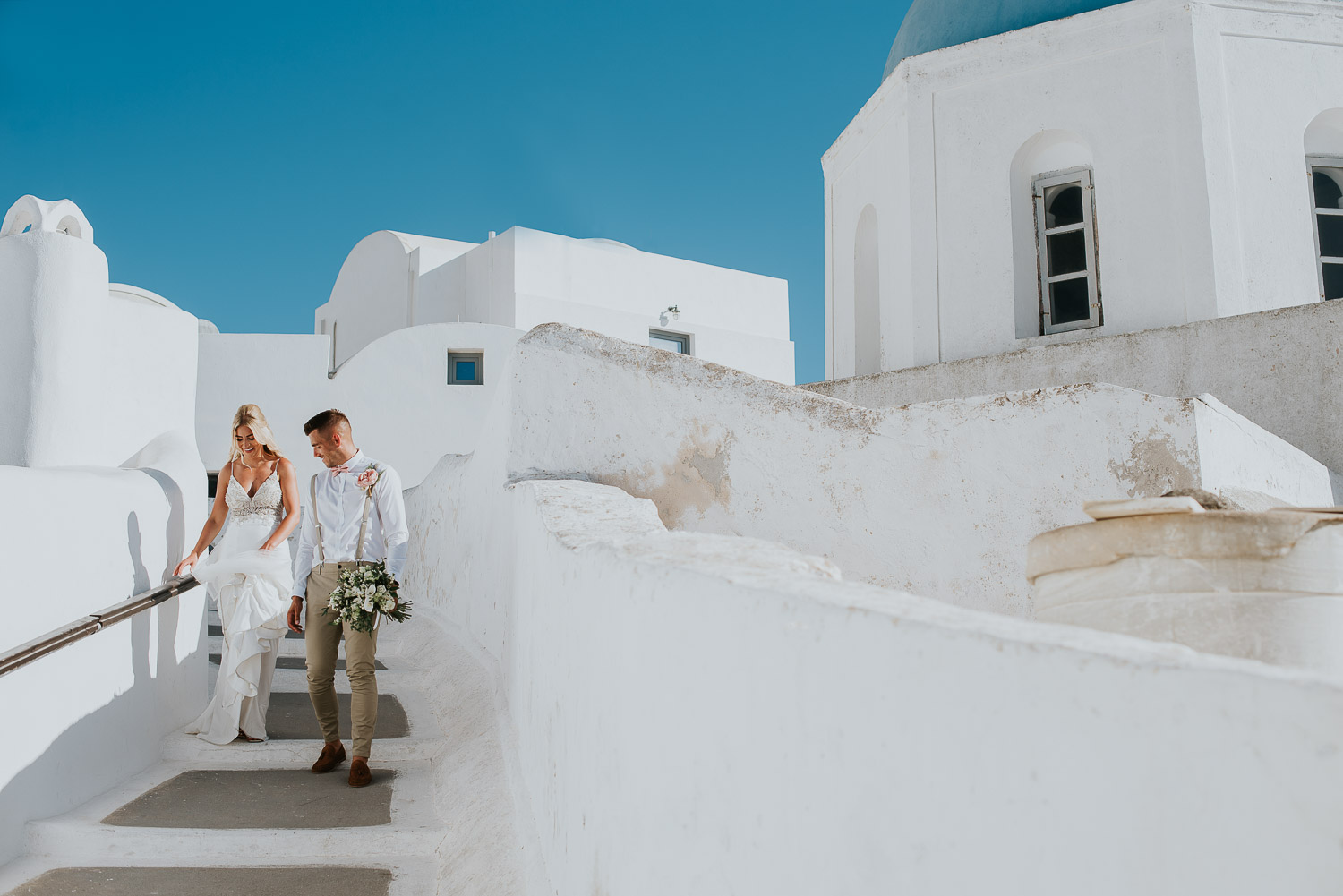 Wedding photographer Santorini: bride and groom walking down the stairs smiling passing blue dome church by Ben and Vesna.
