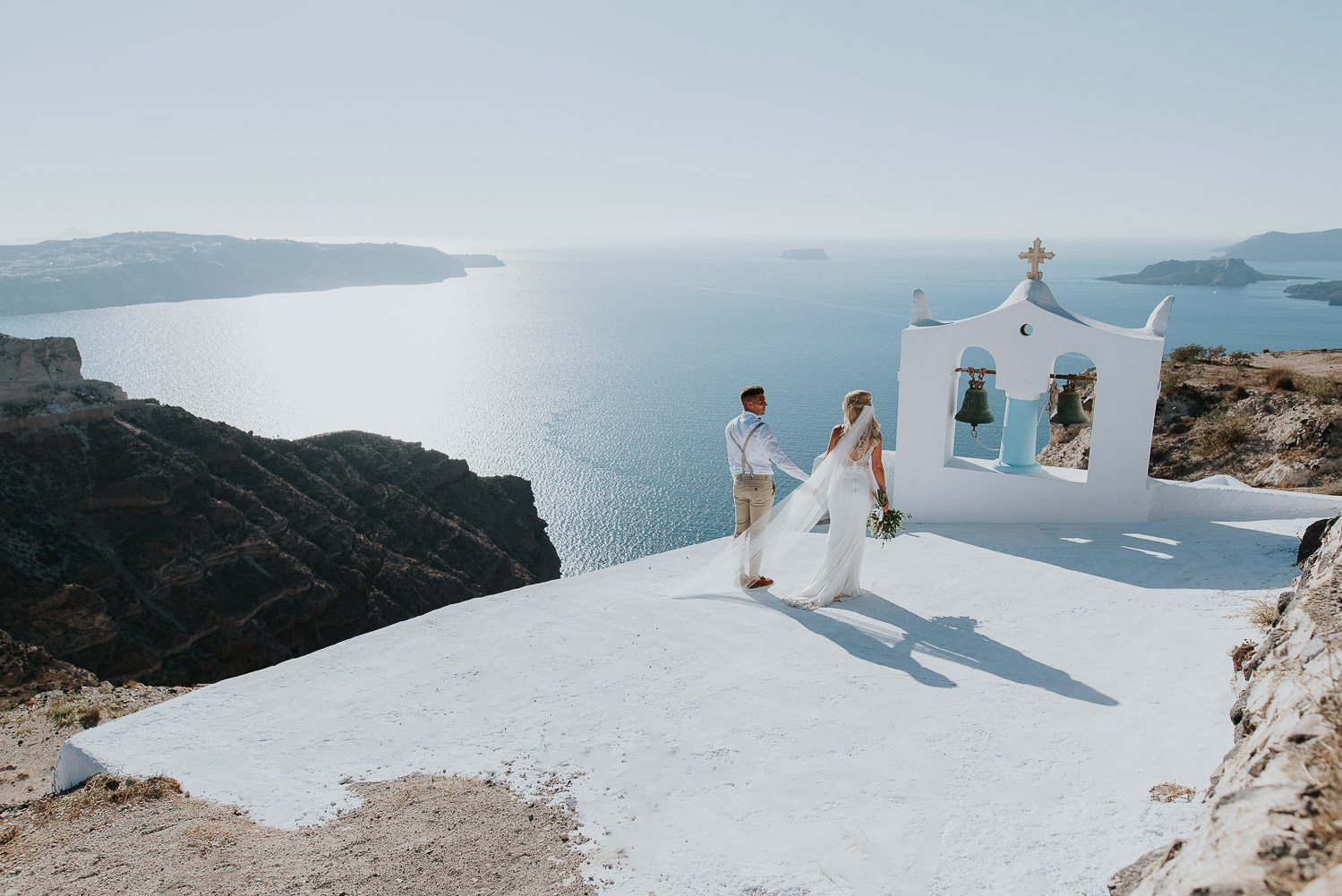 Wedding photographer Santorini: panoramic photo of bride and groom holding hands with bell tower, sea and the sky around them by Ben and Vesna.