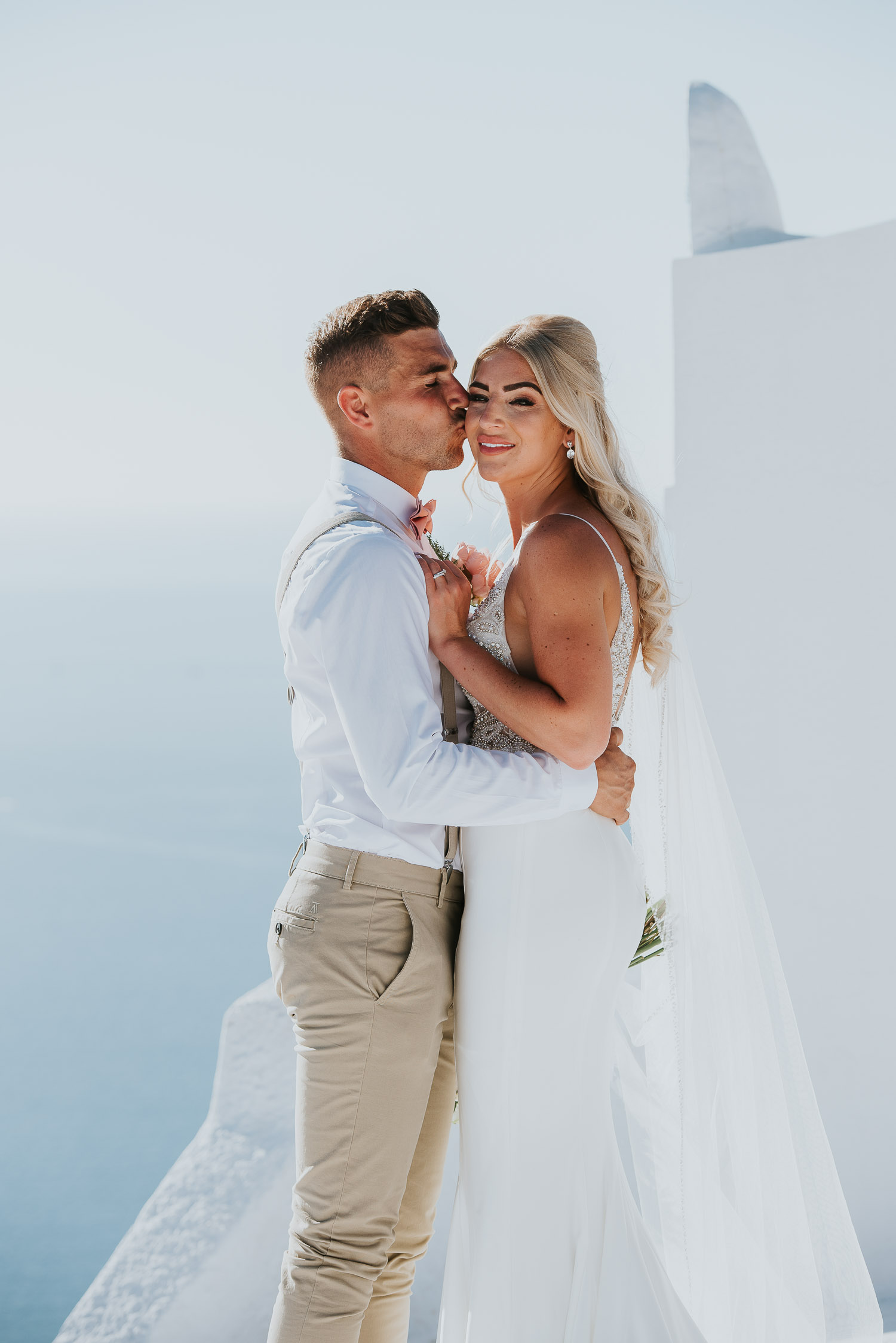 Wedding photographer Santorini: close up of groom kissing bride on a cheek in front of the bell tower basked in the afternoon light by Ben and Vesna.