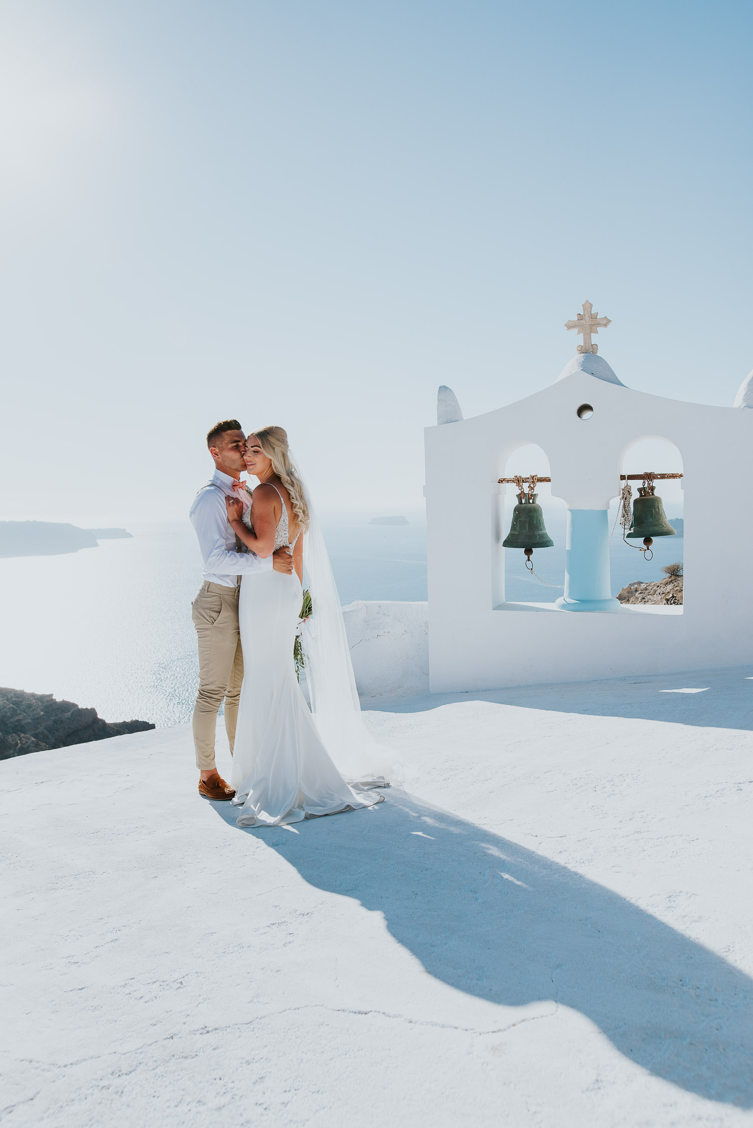 Wedding photographer Santorini: groom kissing bride on a cheek in front of the bell tower basked in the afternoon light by Ben and Vesna.