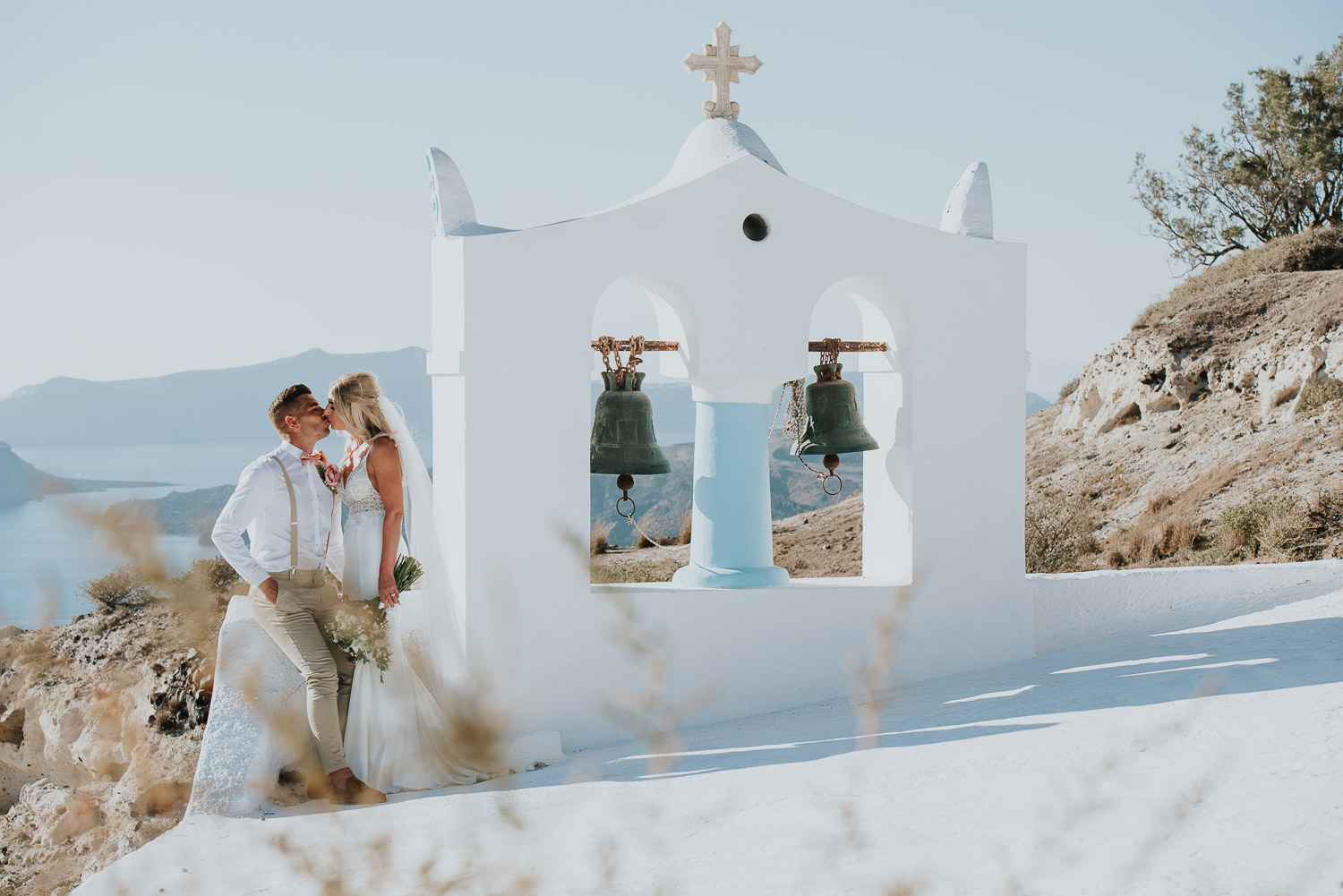 Wedding photographer Santorini: groom kissing bride next to the bell tower basked in the afternoon light by Ben and Vesna.