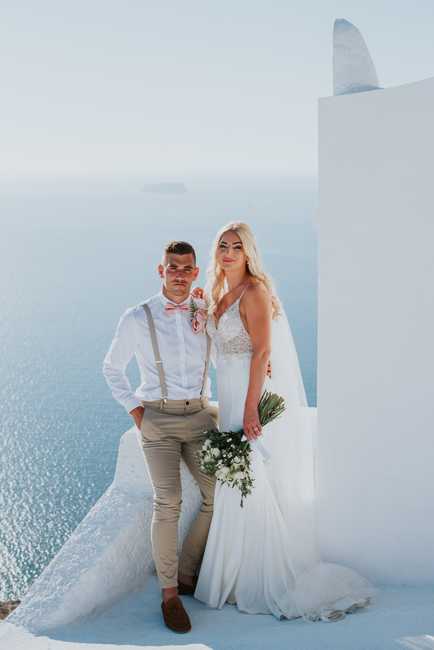 Wedding photographer Santorini: groom and bride looking at the camera next to the bell tower basked in the afternoon light by Ben and Vesna.