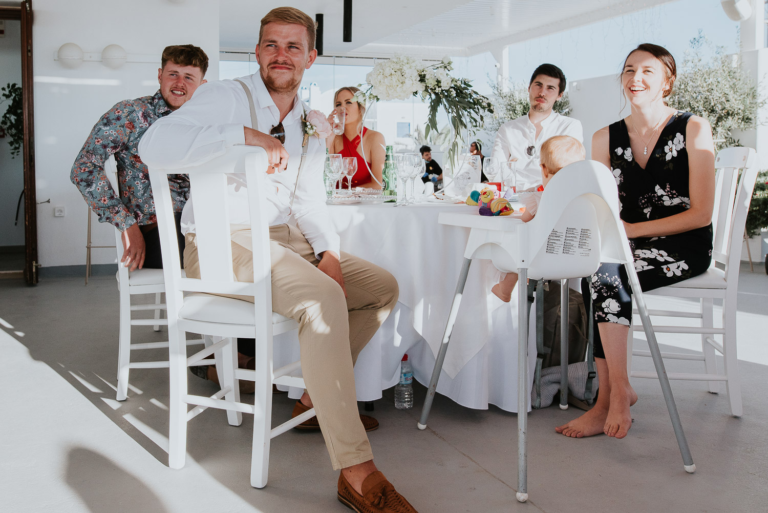 Wedding photographer Santorini: guests sat at their banquet table in the shade smiling as they listen to the speeches by Ben and Vesna.