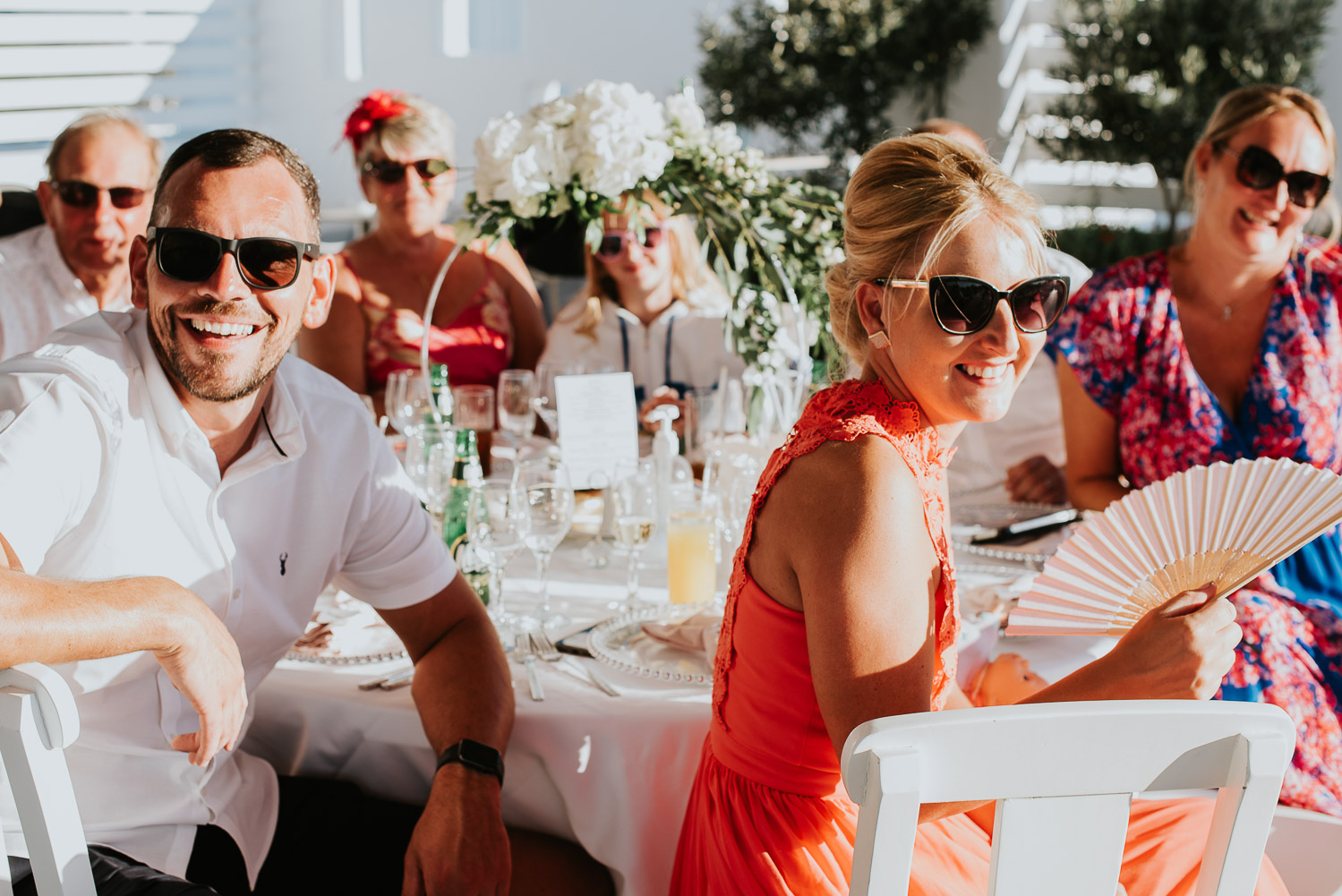 Wedding photographer Santorini: guests sat at their banquet table basked in the sun smiling as they listen to the speeches by Ben and Vesna.