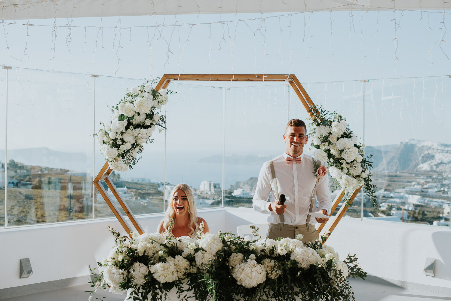 Wedding photographer Santorini: groom and bride in front of the wedding arch basked in the sun as he starts his speech by Ben and Vesna.