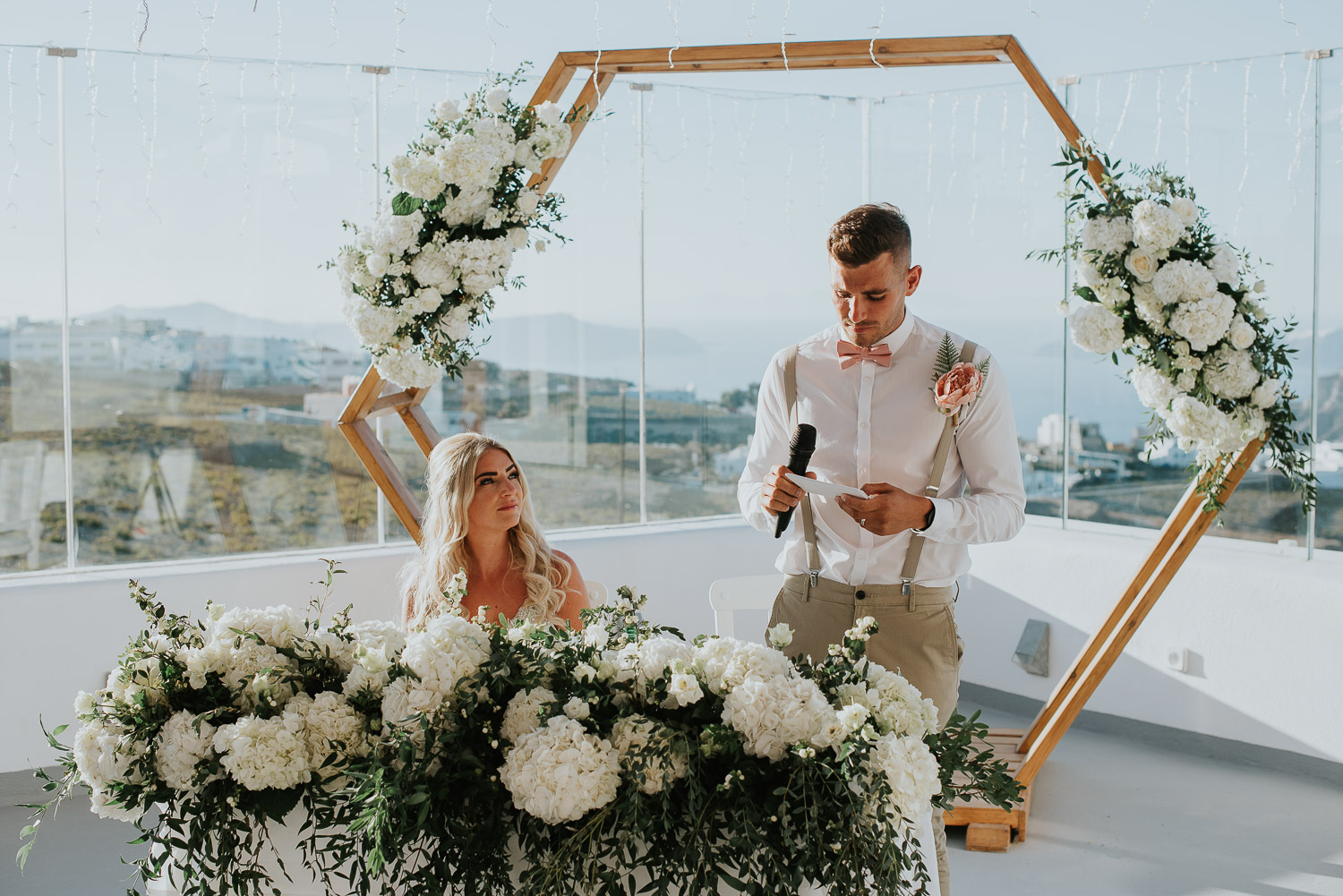 Wedding photographer Santorini: bride looks at her groom as he reads his speech basked in the sun by Ben and Vesna.
