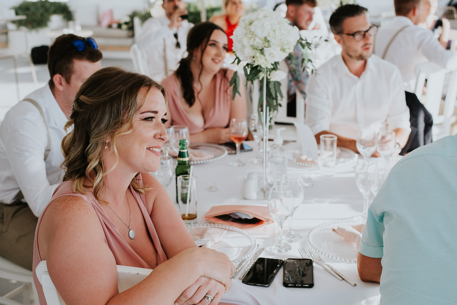 Wedding photographer Santorini: close up of guests sat at their table in the shade smiling as they listen to the speeches by Ben and Vesna.