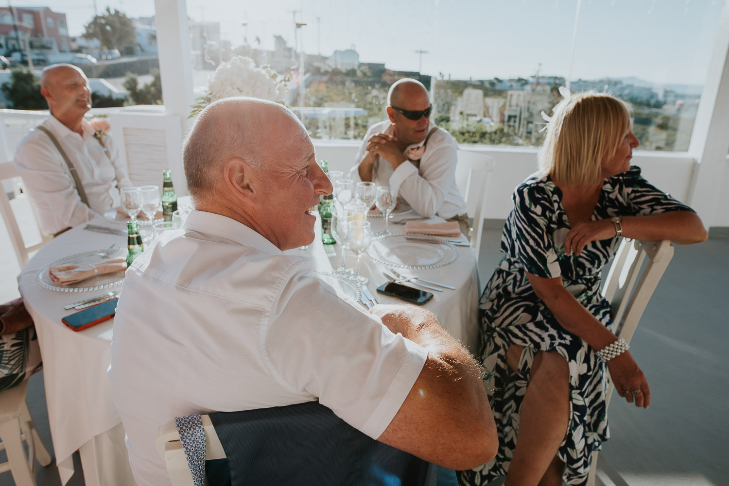 Wedding photographer Santorini: guests sat at their table basked in the sun smiling as they listen to the speeches by Ben and Vesna.