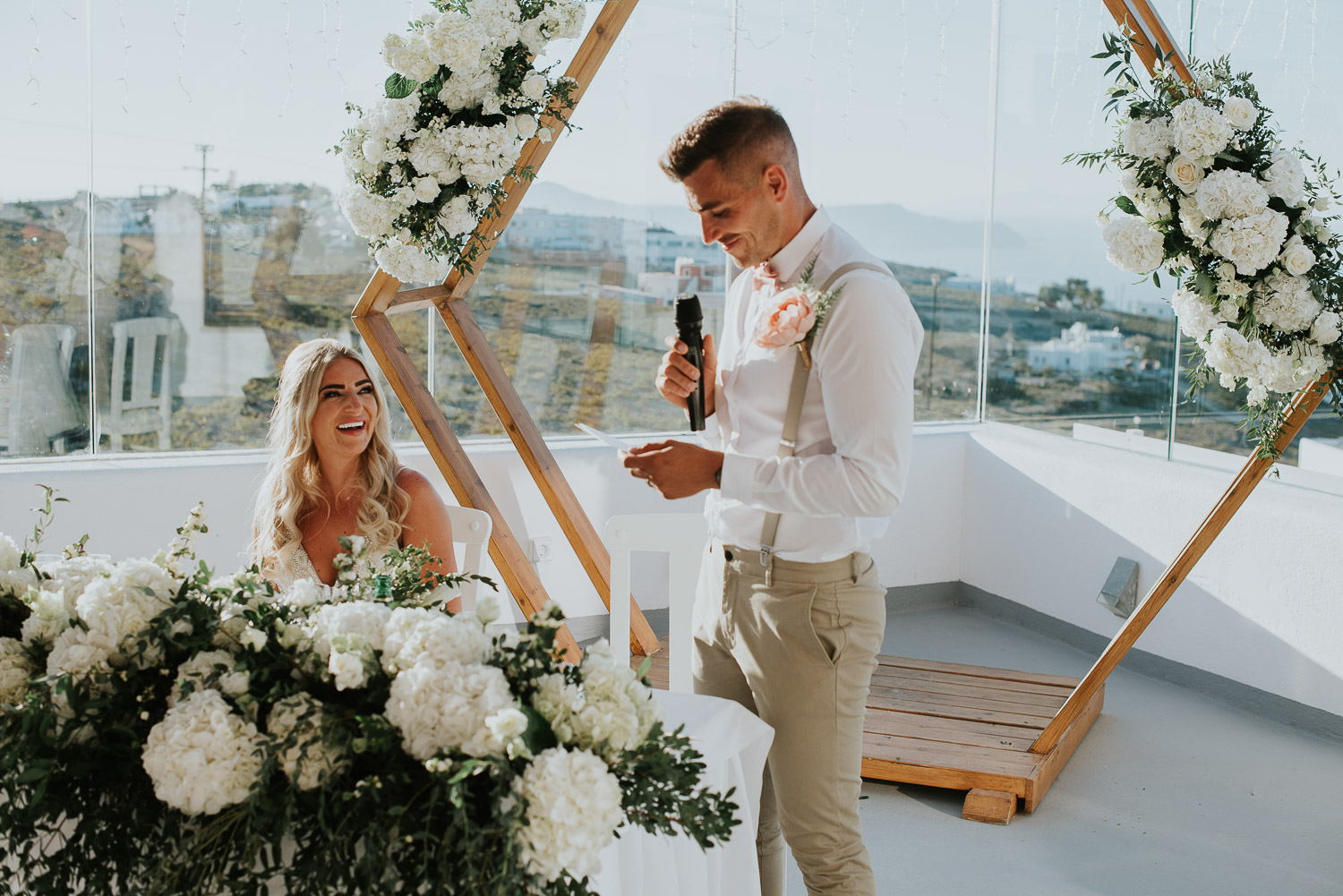 Wedding photographer Santorini: bride smiles as groom reads his speech basked in the sun by Ben and Vesna.