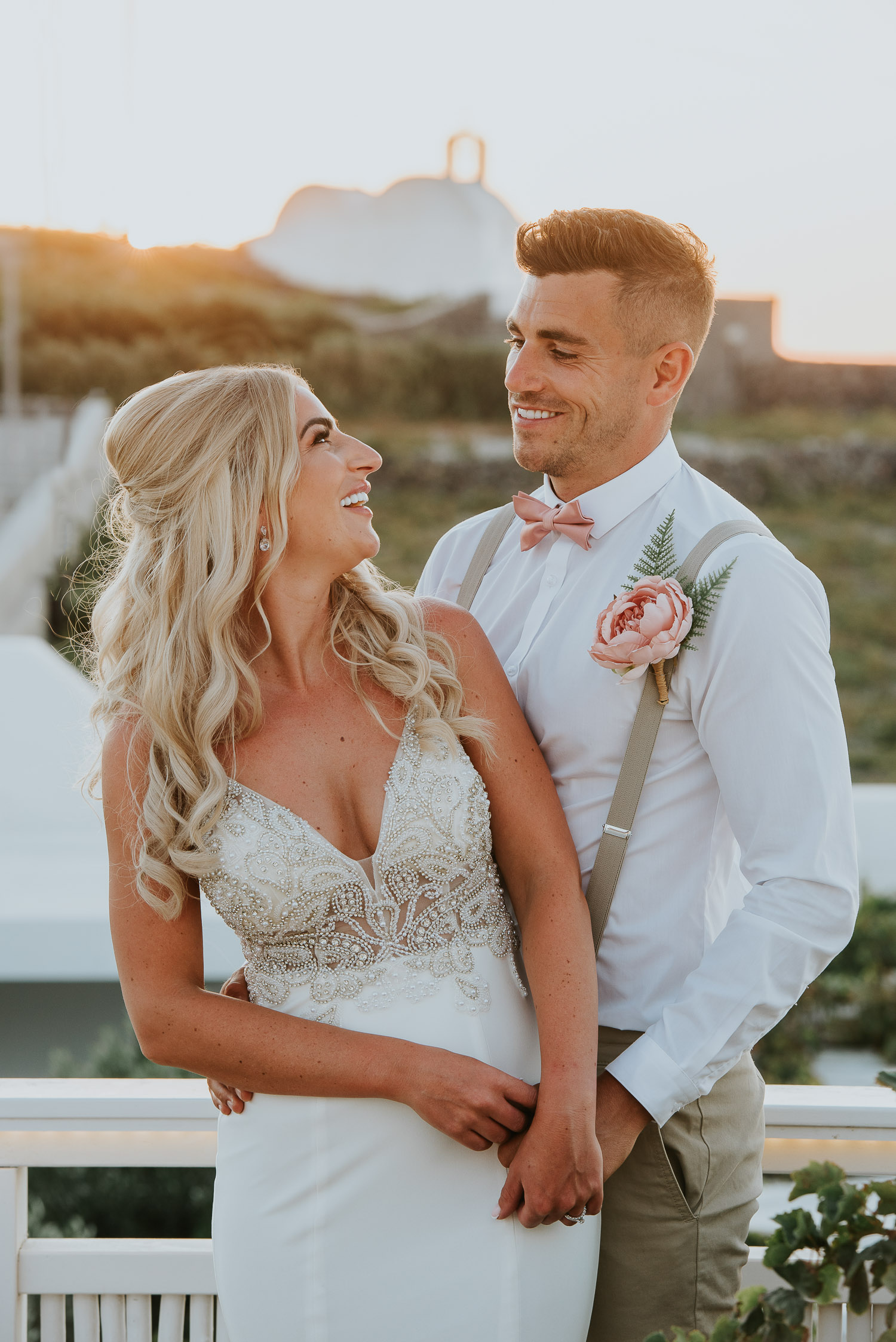 Wedding photographer Santorini: close up of bride and groom smiling backlit by golden sunset light with a church in the background by Ben and Vesna.