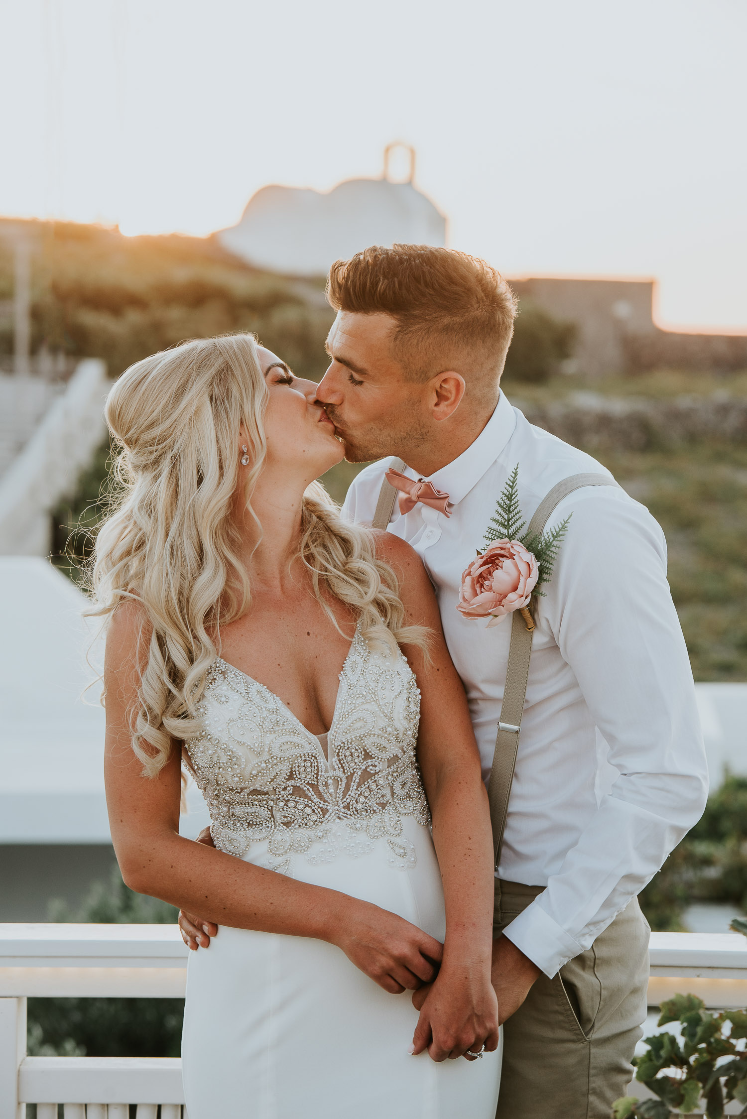 Wedding photographer Santorini: close up of bride and groom kissing backlit by golden sunset light with a church in the background by Ben and Vesna.
