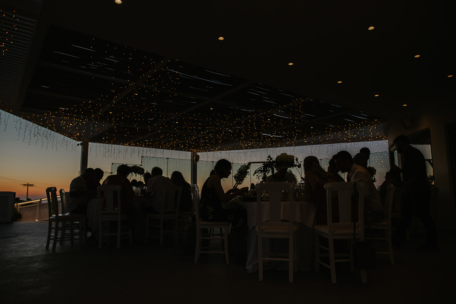 Wedding photographer Santorini: outlines of the guests sat on the reception terrace in the dusk light with fairy lights above them  by Ben and Vesna.