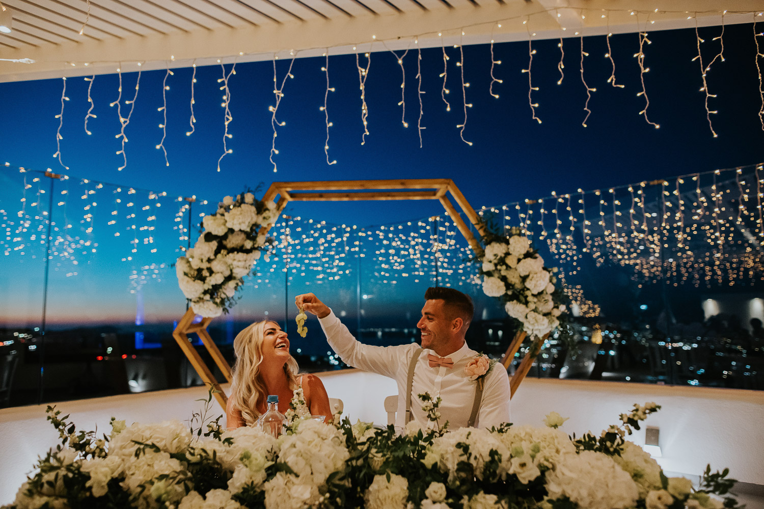 Wedding photographer Santorini: bride and groom laughing as he feeds her grapes surrounded by fairy lights by Ben and Vesna.