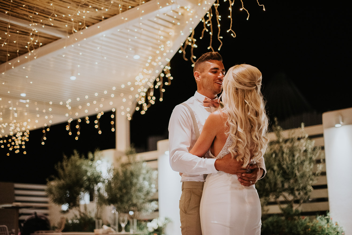 Wedding photographer Santorini: bride and the groom under the fairy lights embraced for the first dance by Ben and Vesna.