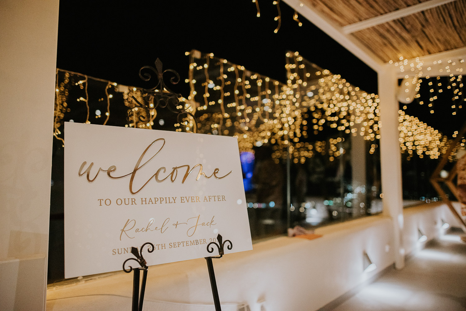 Wedding photographer Santorini: welcome sign beautifully lit by the fairy lights reflecting in the glass wall by Ben and Vesna.
